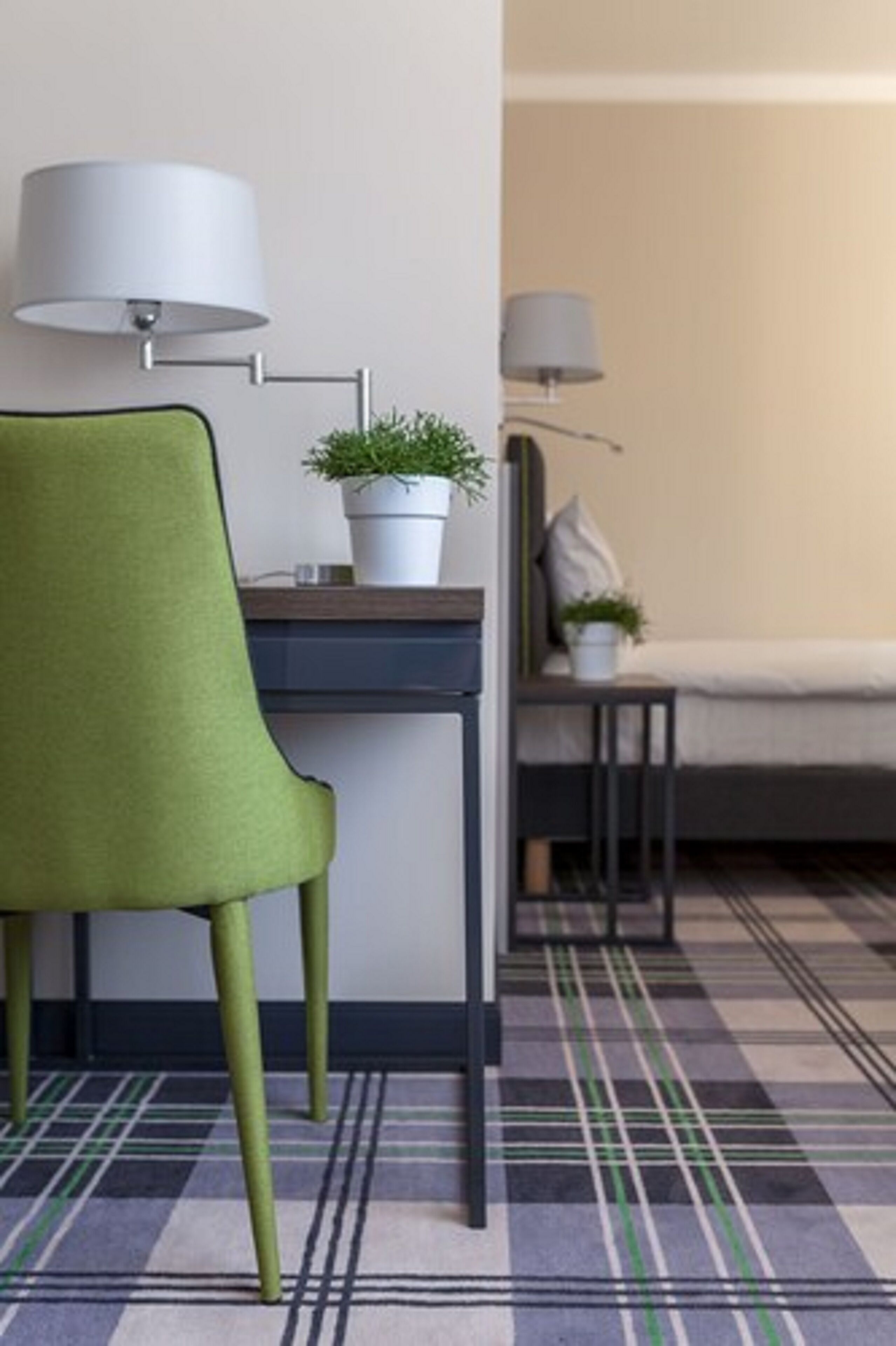 Una habitació amb una cadira verda, un escriptori amb una planta en test i un llit amb un almoiner.