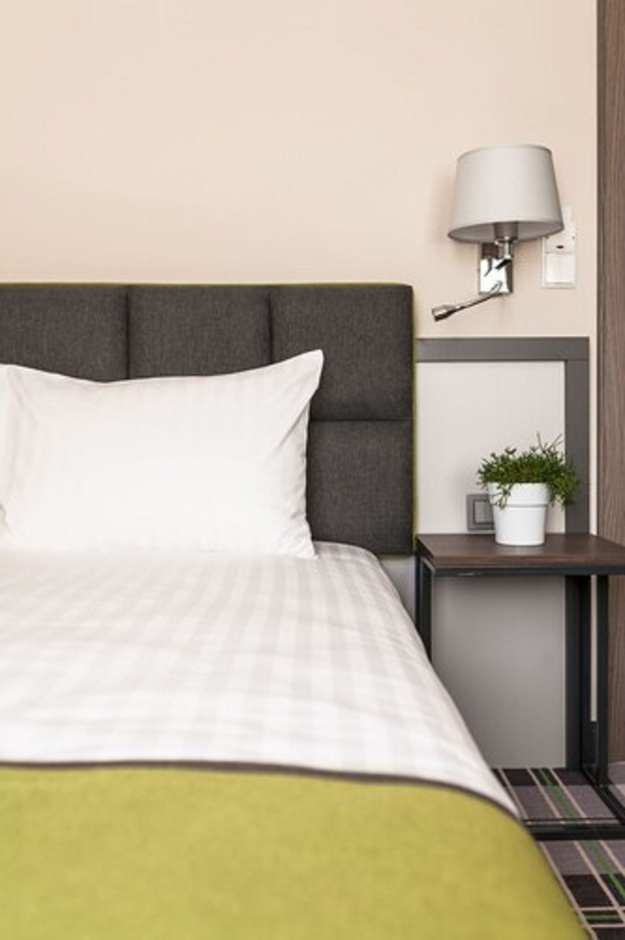Una cama bien hecha con cabecera gris, una mesita de noche con lámpara y una pequeña planta en maceta.