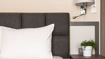 Una cama bien hecha con cabecera gris, una mesita de noche con lámpara y una pequeña planta en maceta.