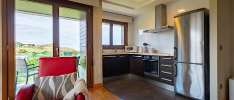 A modern kitchen with a stainless steel refrigerator, a microwave, and a built-in oven. There is a red chair with a patterned cushion in the living area, and large windows offering a view of the outdoors.