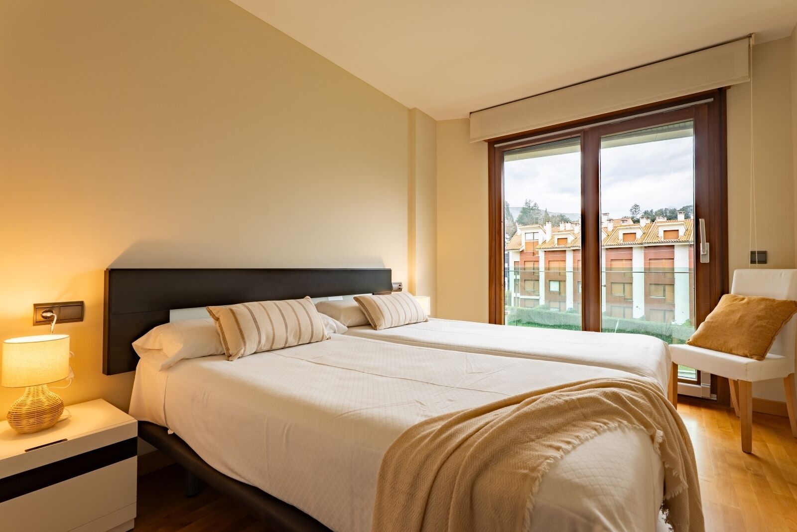Habitación de hotel con una cama grande, una mesita de noche con lámpara, una silla y vistas a edificios a través de un ventanal amplio.