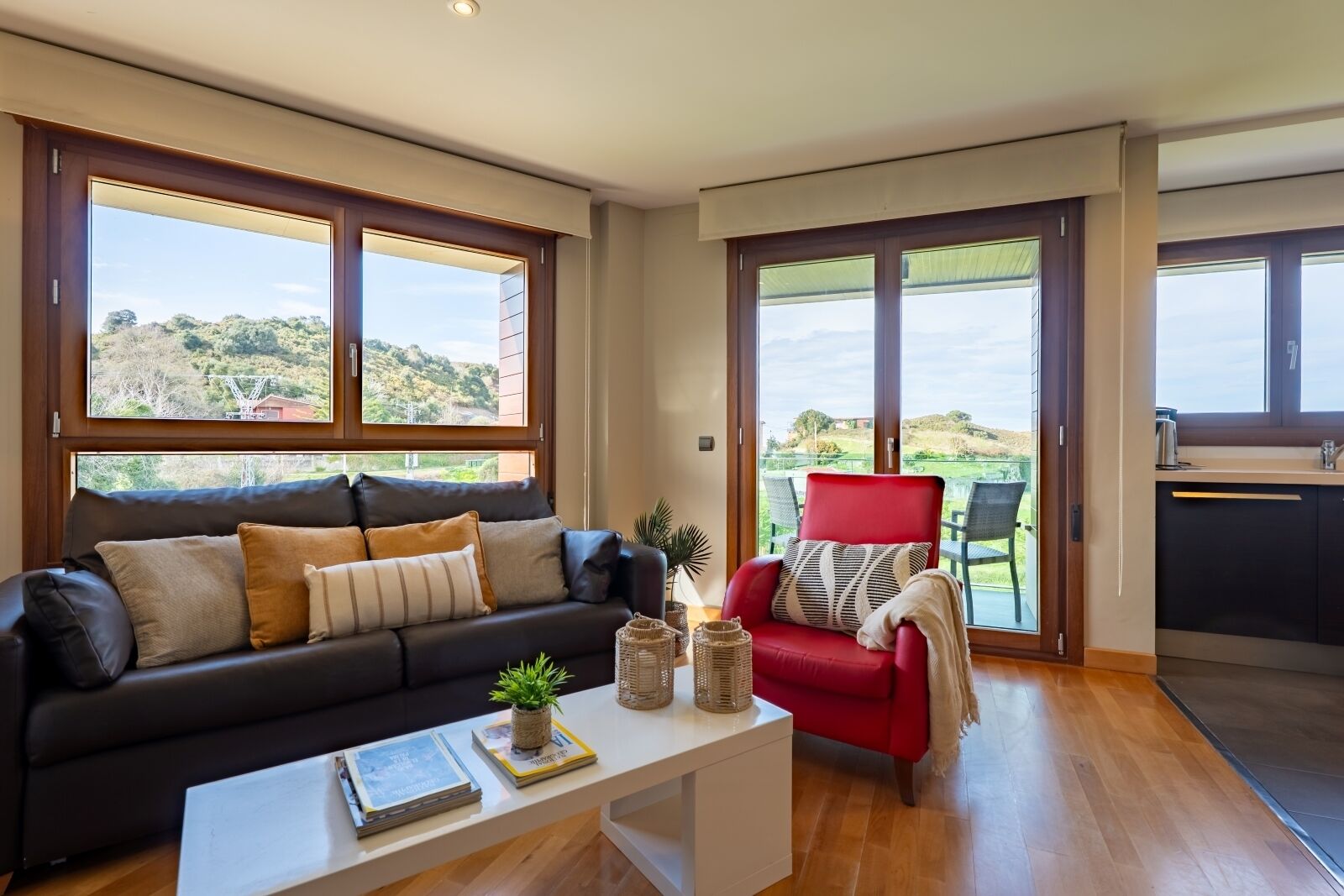 Ein modernes Wohnzimmer mit einer schwarzen Ledersofa, einem roten Sessel und einem weißen Couchtisch. Große Fenster bieten Blick auf eine grüne Landschaft.