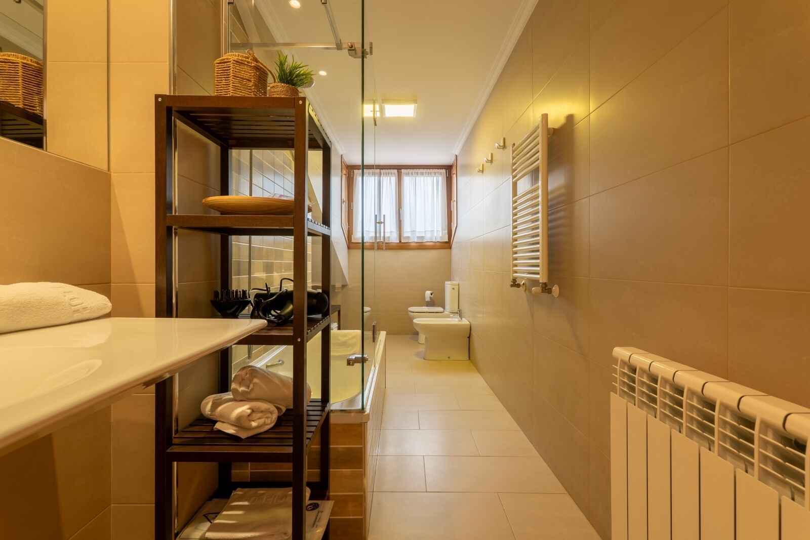 Baño estrecho con lavamanos blanco, inodoro y radiador. Se observa una estantería con toallas y una cesta en la pared.