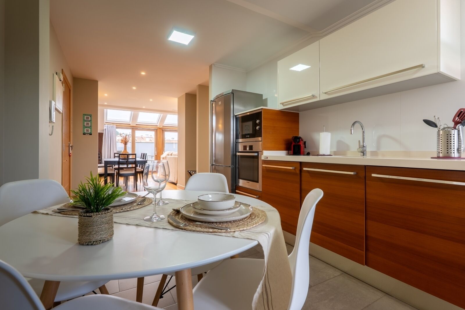 Une cuisine moderne avec des meubles en bois, un réfrigérateur en acier inoxydable et un coin repas avec une table blanche mise pour deux personnes.