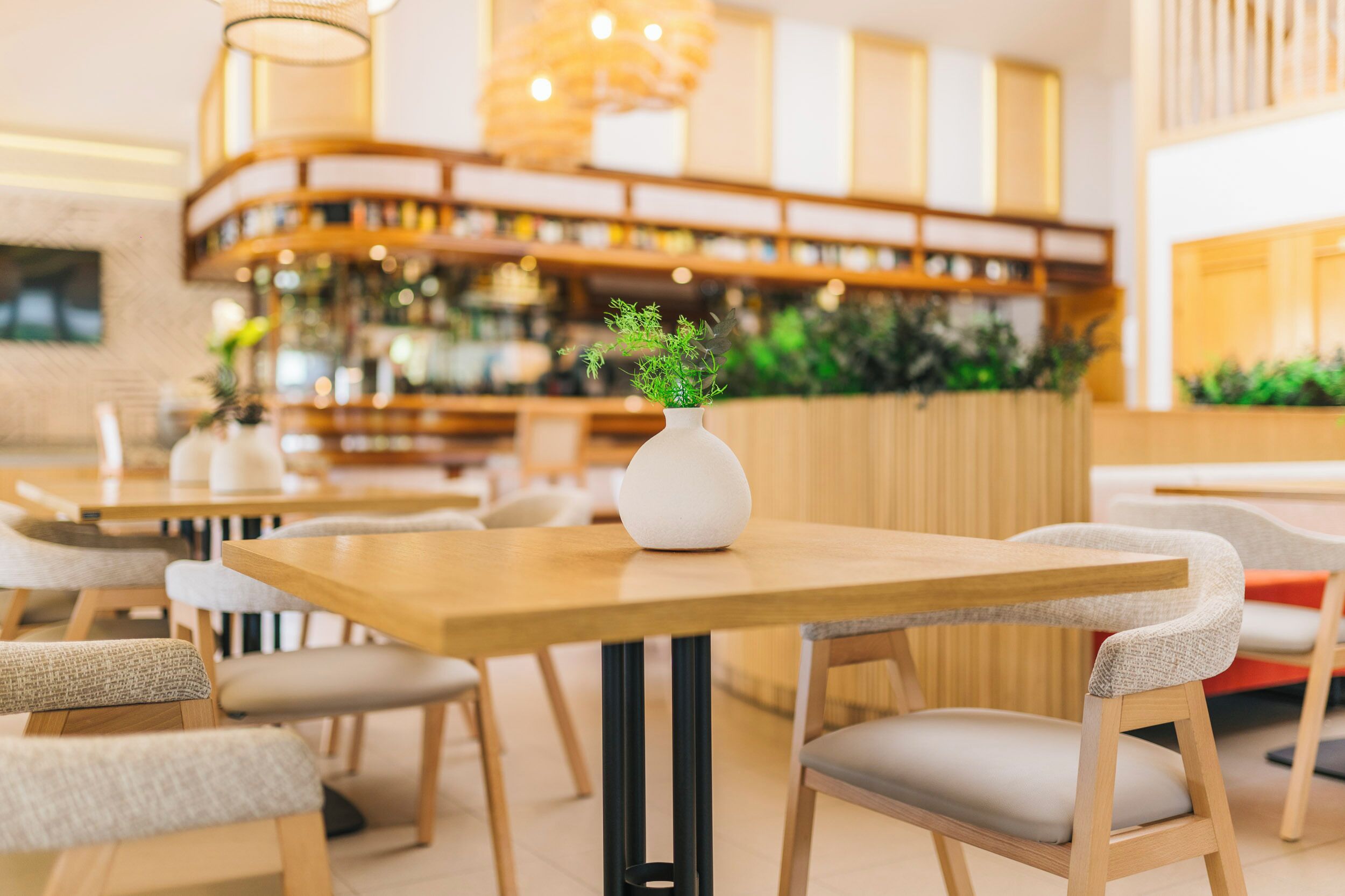 Un comedor moderno con mesas y sillas de madera, un florero blanco con plantas, y una barra en el fondo.