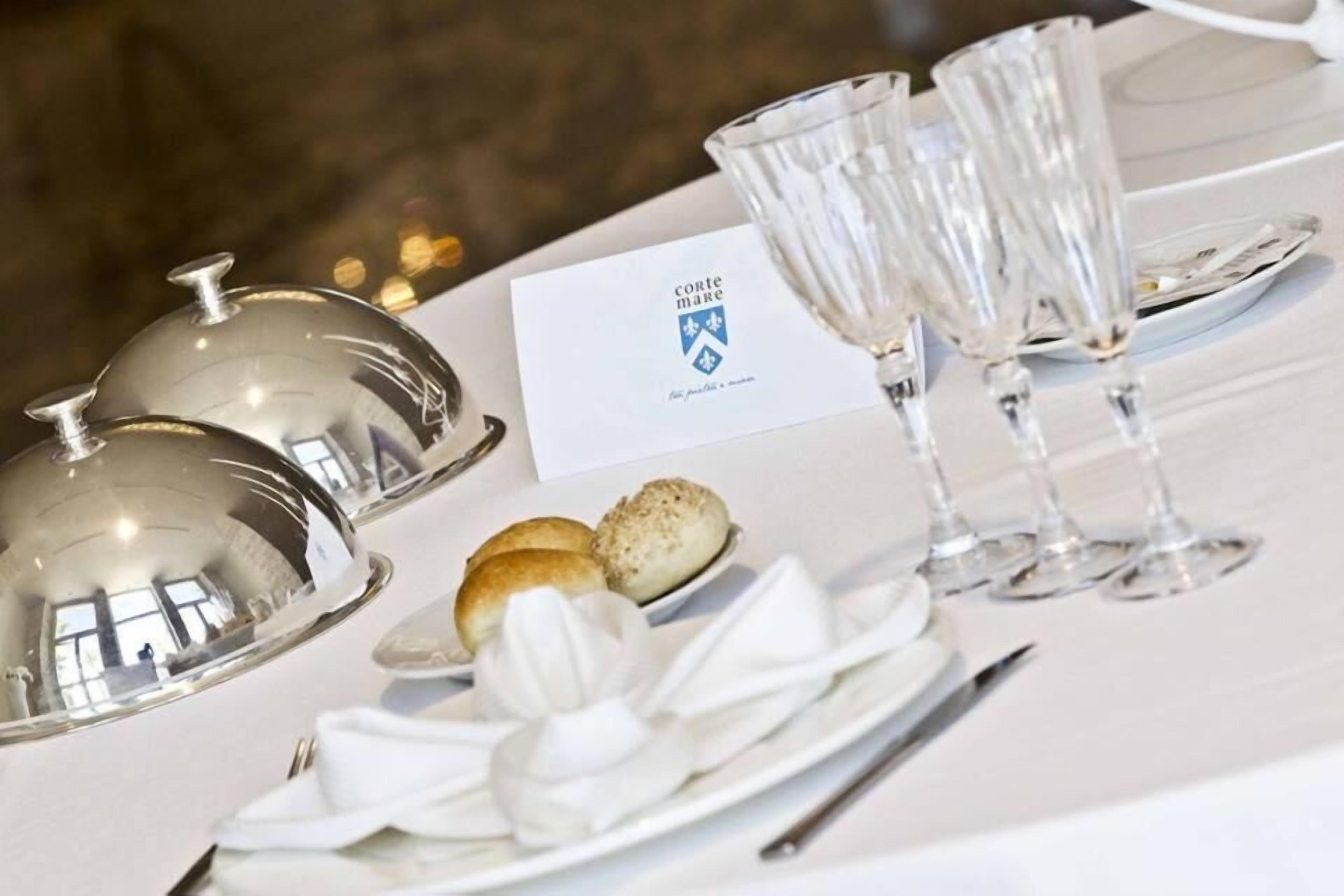 A table set with a white tablecloth, a silver cloche, a glass, a plate with a dessert, and a card with a crest.
