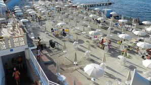 Zona junto a la playa con numerosas sombrillas blancas y tumbonas, un muelle que se adentra en el mar y una piscina con gente.