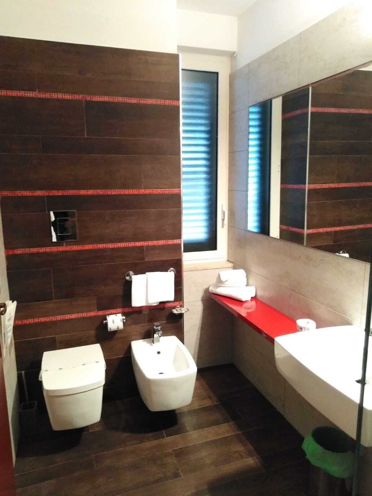 A modern bathroom with dark wood flooring, a white toilet, a bidet, a red countertop, and a large mirror.