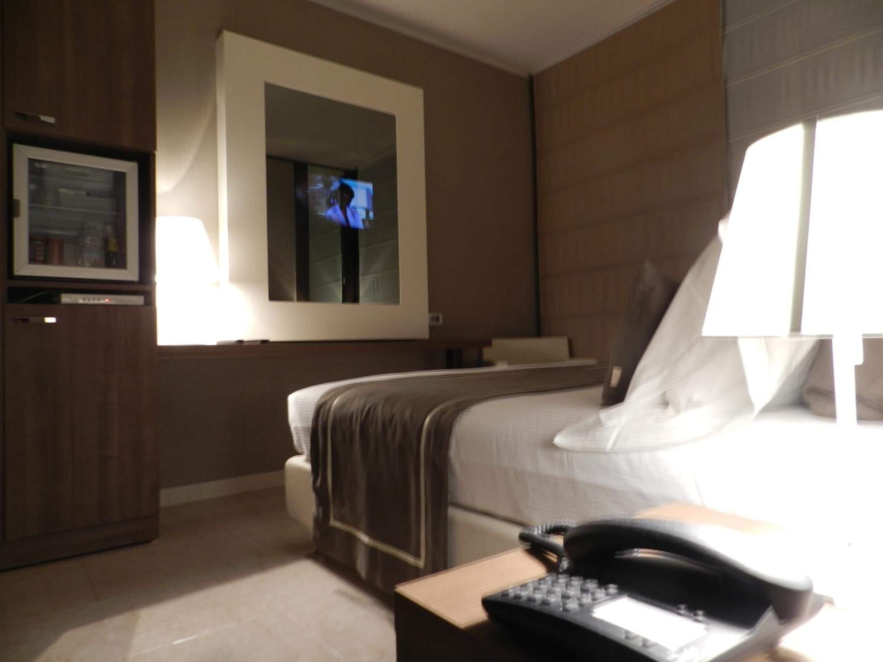 A hotel room with a bed, a desk with a telephone, a television, and a mirror on the wall.