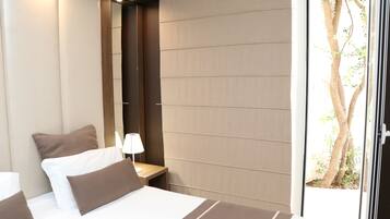 Una habitación de hotel moderna con una cama grande, una mesita de noche, una lámpara y un ventanal corredero que da a un balcón con un árbol.