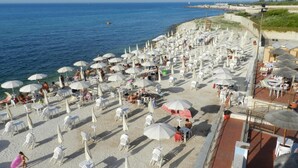 Una playa con numerosas sombrillas y sillas blancas, vista despejada al mar y un paseo marítimo con barandilla.