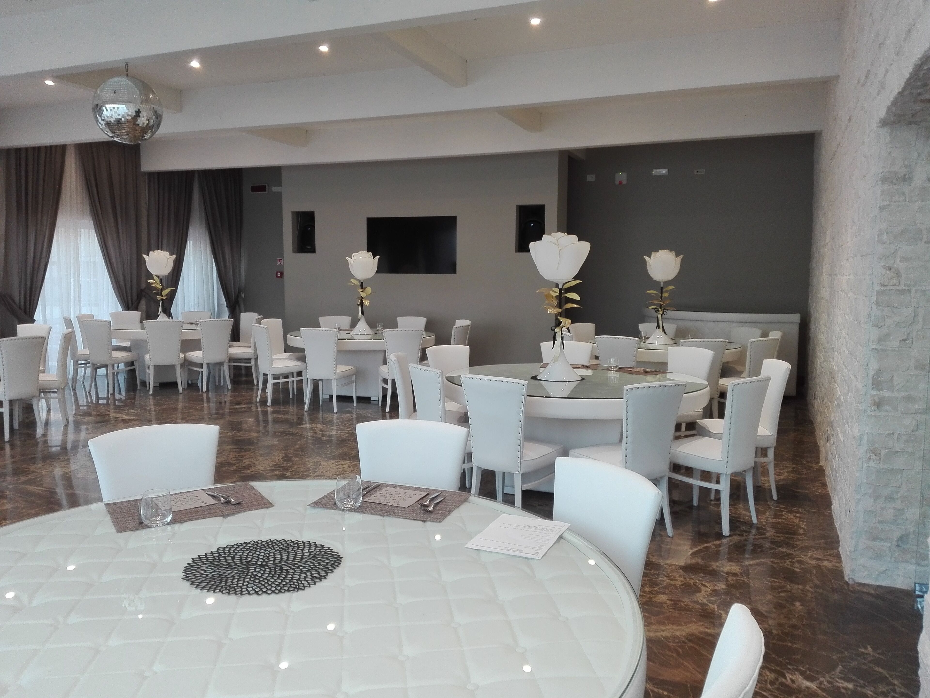 Un comedor amplio y moderno con mesas redondas y sillas blancas, con elegantes centros de mesa florales y una bola de discoteca.