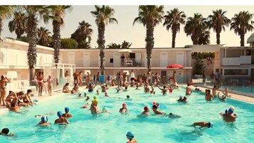 Una piscina concurrida, gente disfrutando del agua, rodeada de palmeras y un edificio con varios balcones.