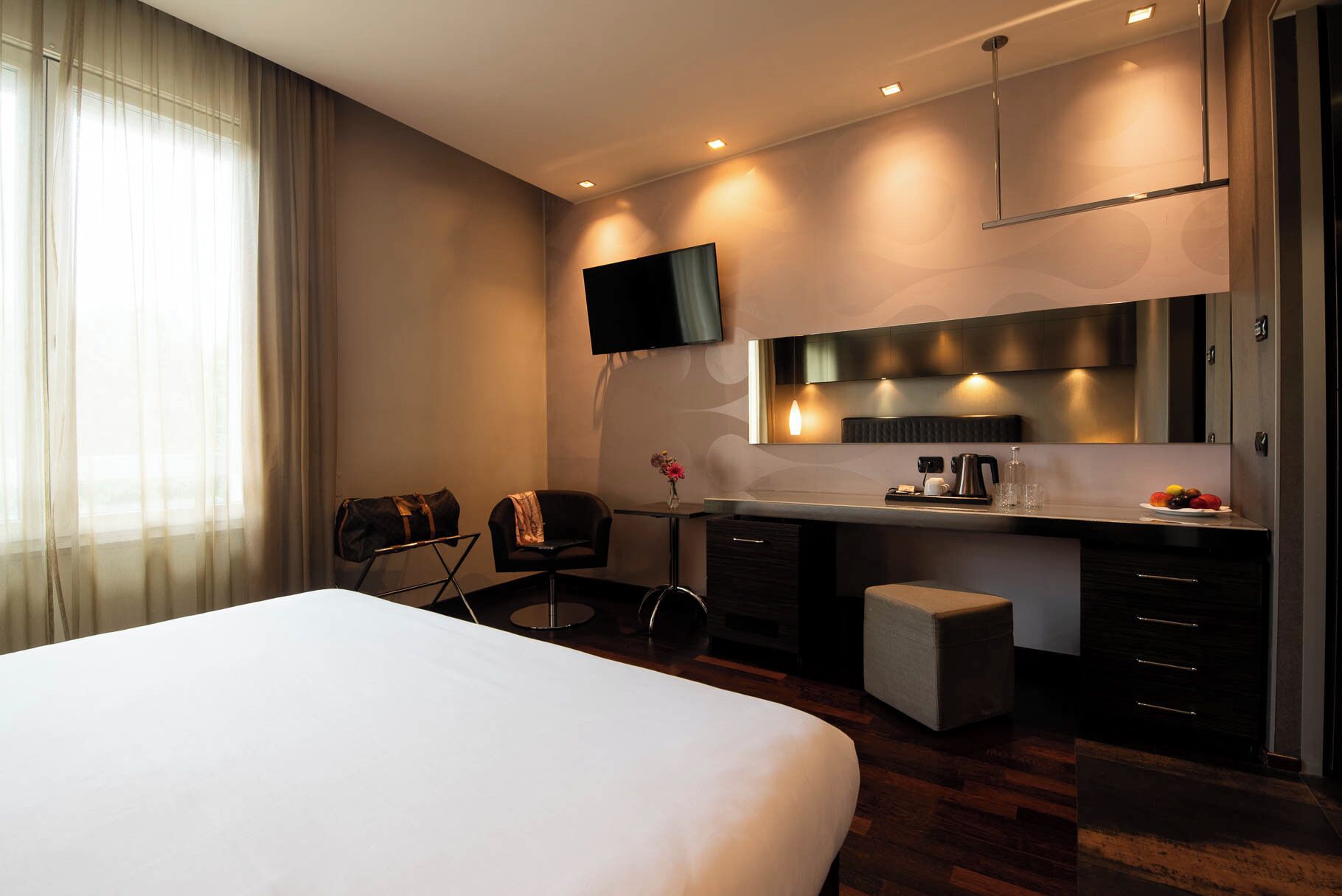 Une chambre d’hôtel moderne dotée d’un grand lit, d’un bureau avec une chaise, d’une télévision à écran plat, d’un miroir et d’une petite table sur laquelle est posé un vase de fleurs.