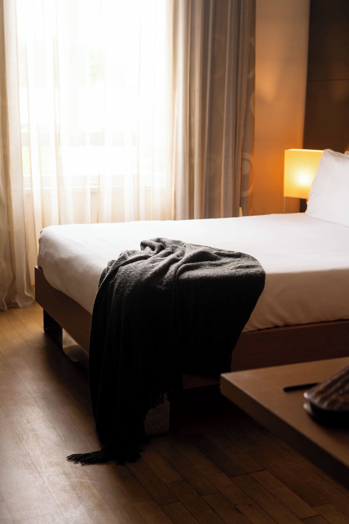 A hotel room with a bed, a bedside table, a lamp, and a window with curtains.