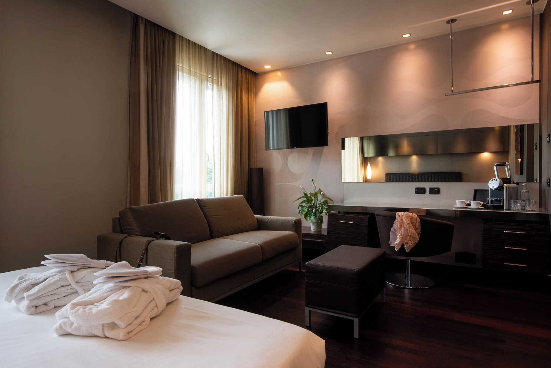A modern hotel room with a large bed, a sofa, a desk with a chair, a television, and a small kitchenette.