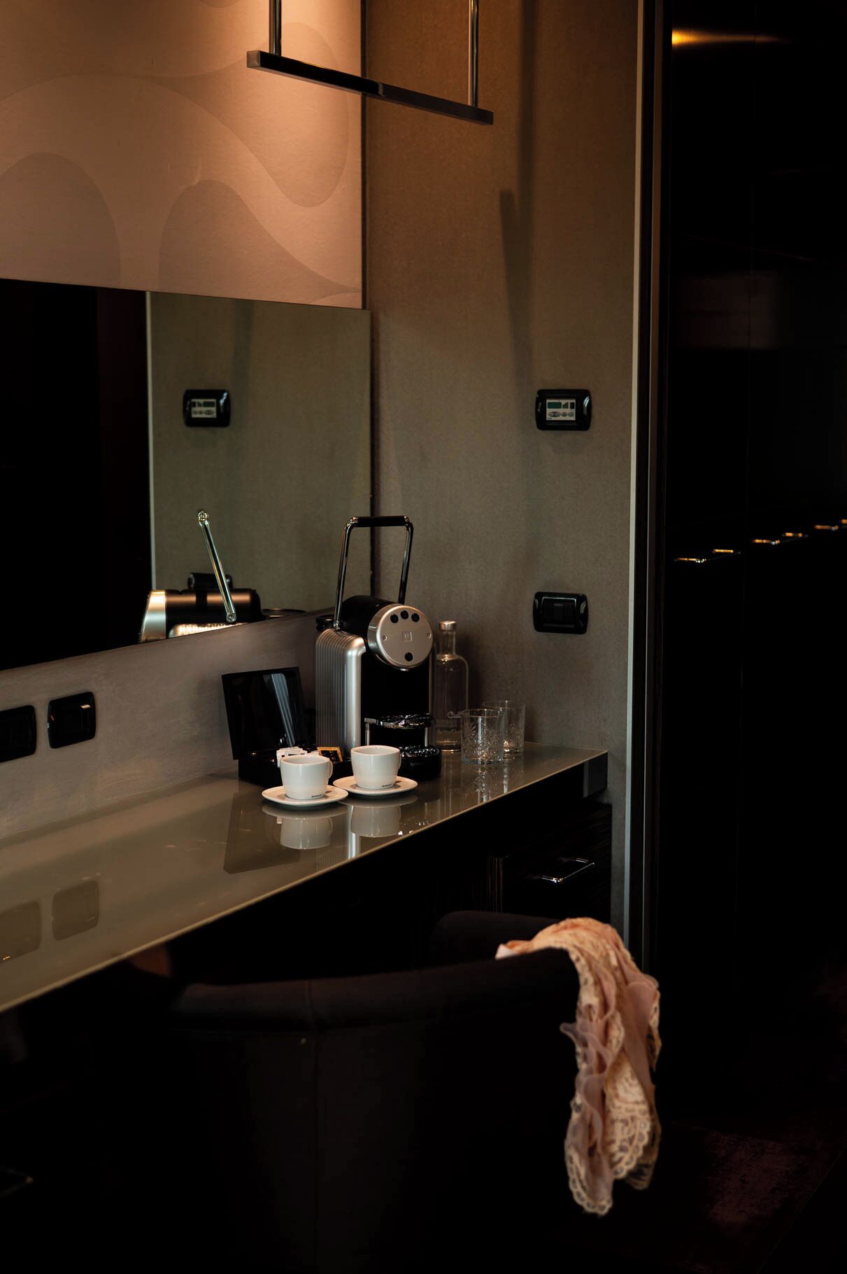 A modern hotel room with a minimalist design, featuring a sleek coffee machine, two cups, a glass, and a towel on a chair.