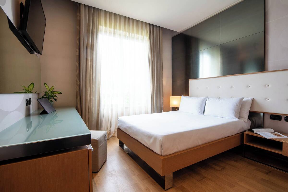 A hotel room with a bed, bedside table, lamp, and a desk with a plant.