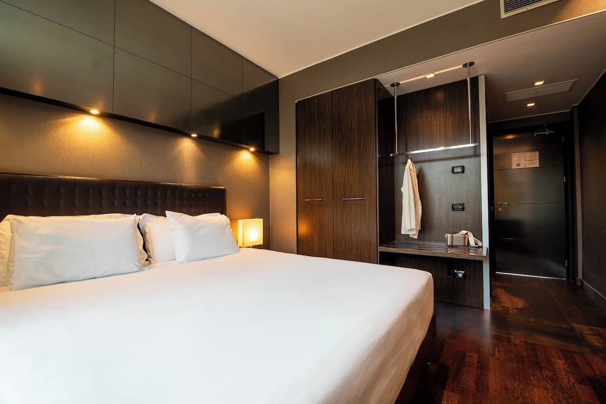 A modern hotel room with a large bed, dark wood wardrobe, and a built-in bench with a towel hanging on it.