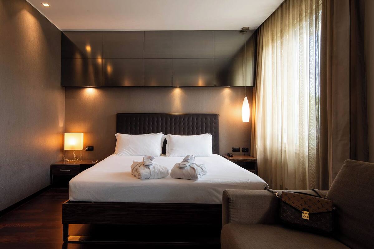 A hotel room with a large bed, a nightstand with a lamp, a chair, and a window with curtains.