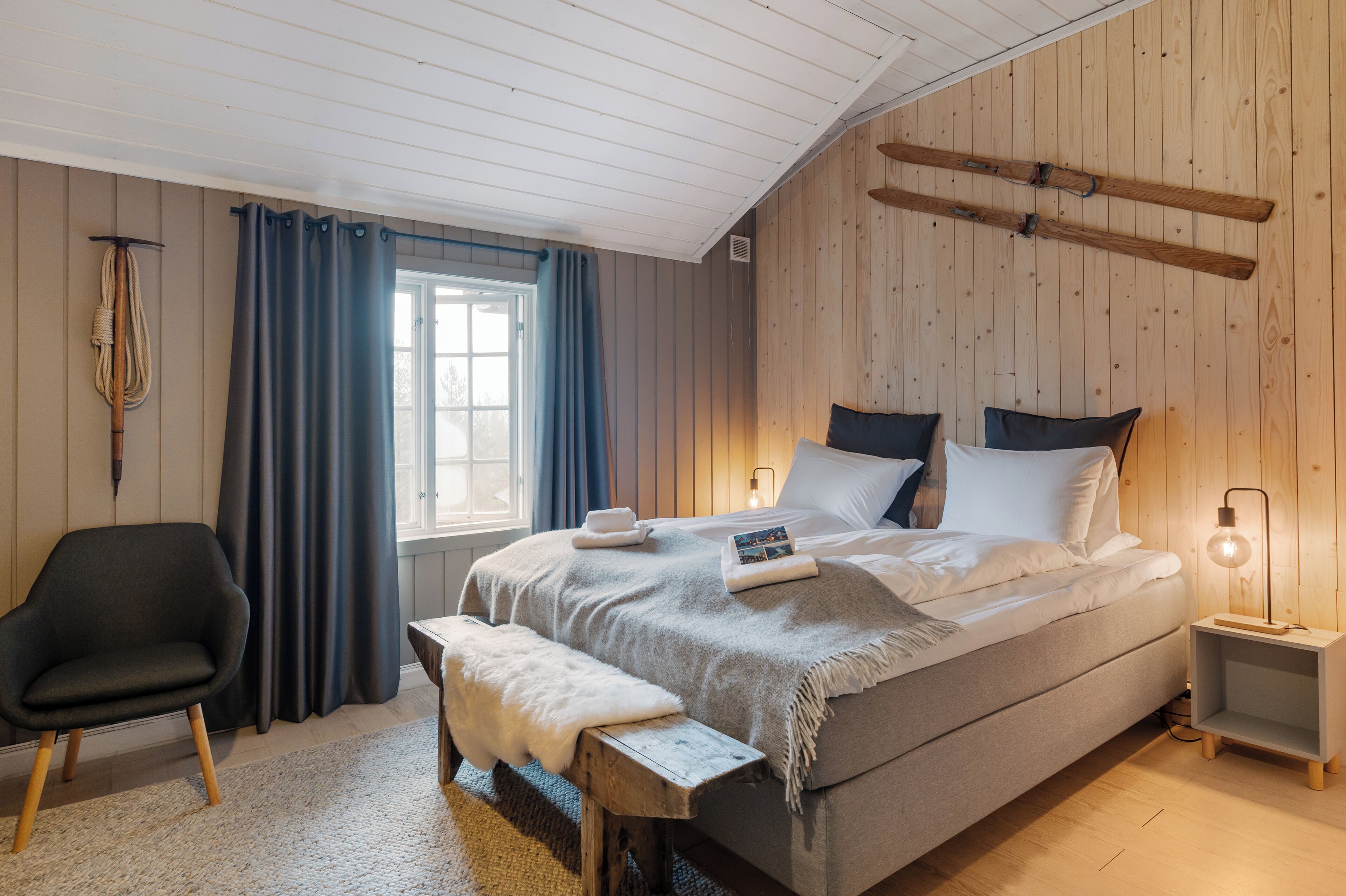 A bedroom with a large bed, a bench, a chair, a bedside table, and a window with curtains.