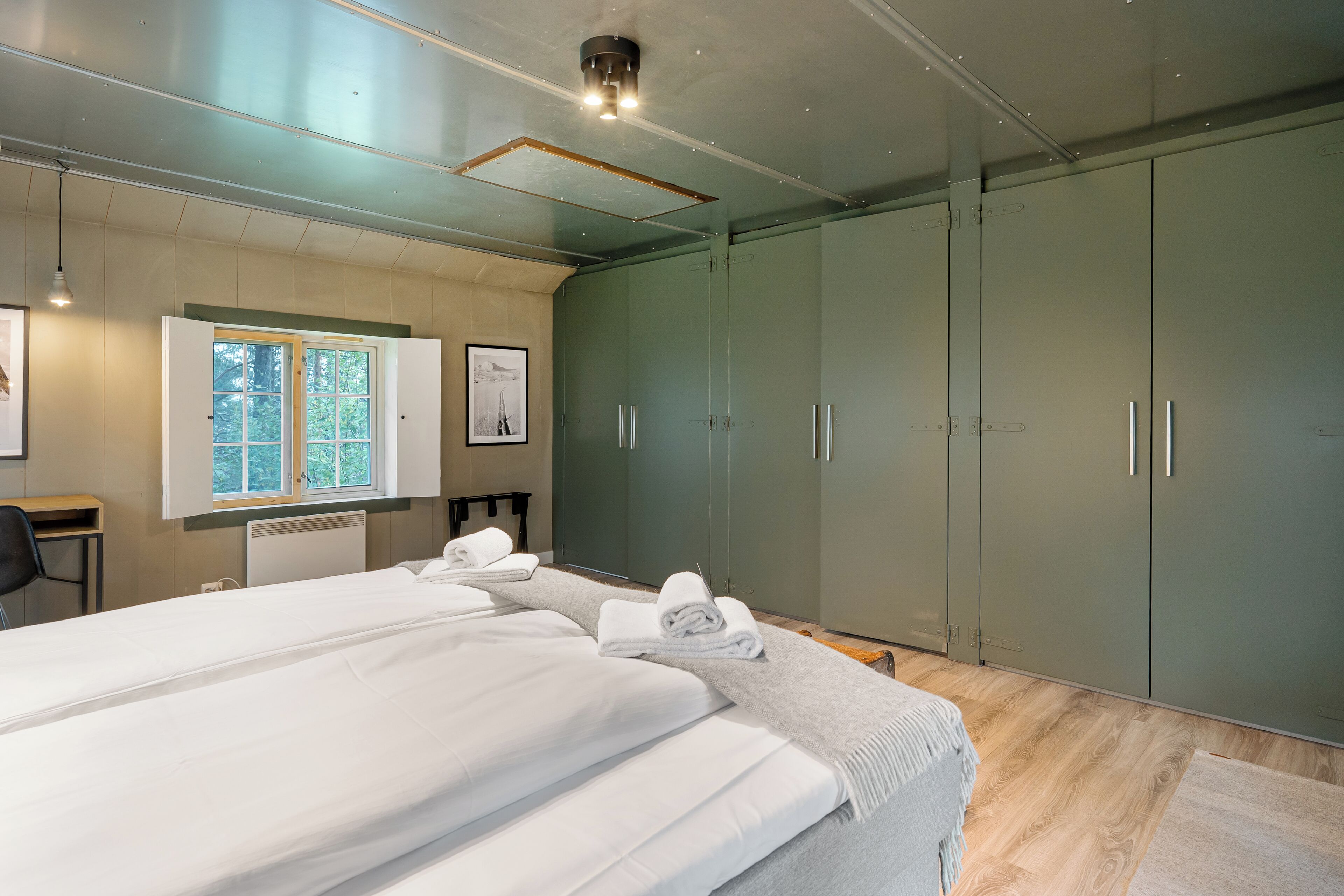 A modern bedroom with two beds, a large wardrobe, a desk, and a window with a view of trees.