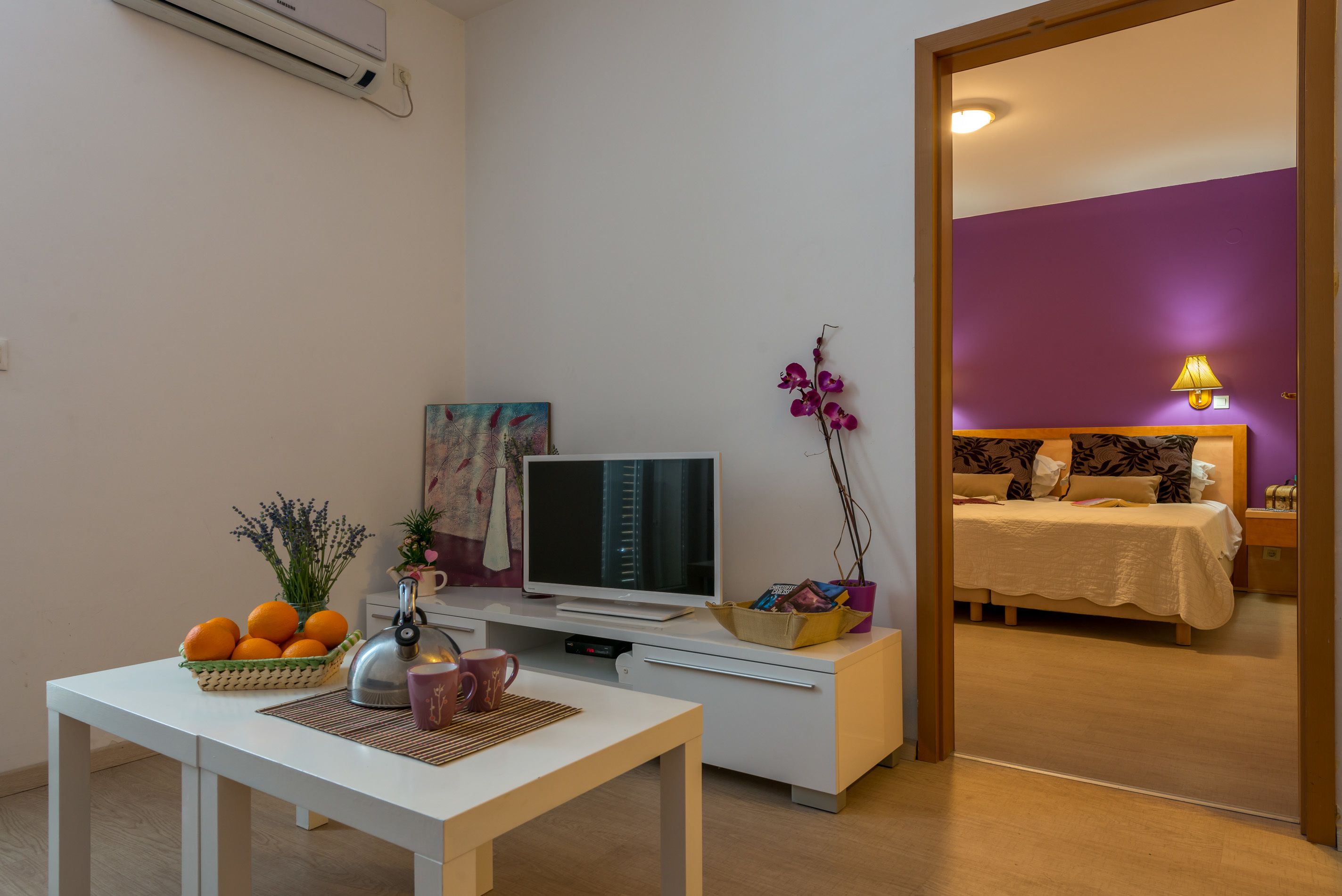 A modern living room with a flat-screen TV, a dining table with a basket of oranges, a coffee maker, and a decorative orchid.