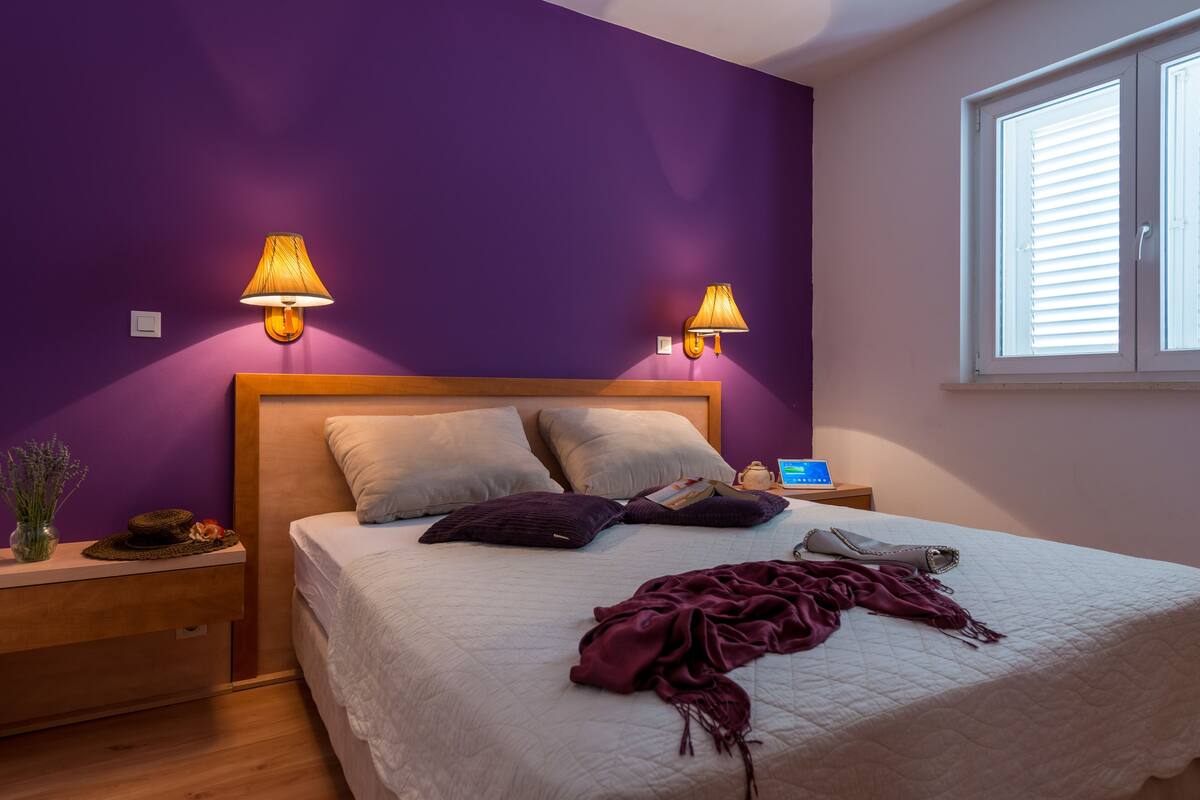 Un dormitorio con paredes moradas, cabecera de madera, dos lámparas de noche, una mesita de noche con lámpara y una ventana con persianas. La cama tiene un edredón blanco y almohadas.