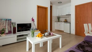 A modern living room with a flat-screen TV, a white coffee table with a fruit basket, a kettle, and a vase with flowers. - Apartments Lapad (Dubrovnik)