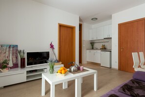 Una sala de estar moderna con un televisor de pantalla plana, una mesa de centro blanca con una cesta de frutas, una tetera y un florero con flores.