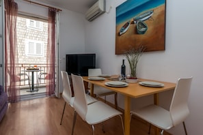 A dining area with a wooden table, white chairs, a painting of boats, and a TV on the wall. - Apartments Lapad (Dubrovnik)