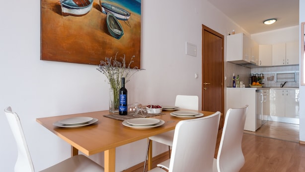 A dining area with a wooden table, white chairs, and a painting of boats on the wall. - Apartments Lapad (Dubrovnik)