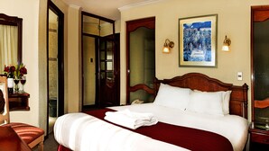 Quarto de hotel com uma cama grande, cabeceira de madeira, mesa de cabeceira com abajur, televisão e um quadro na parede.