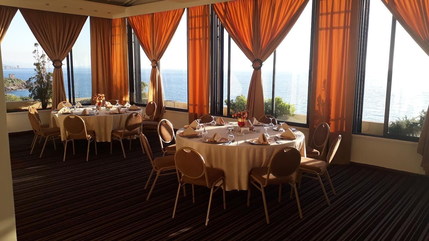 Un comedor con mesas redondas dispuestas para cuatro, ventanas amplias con cortinas y vista al mar.