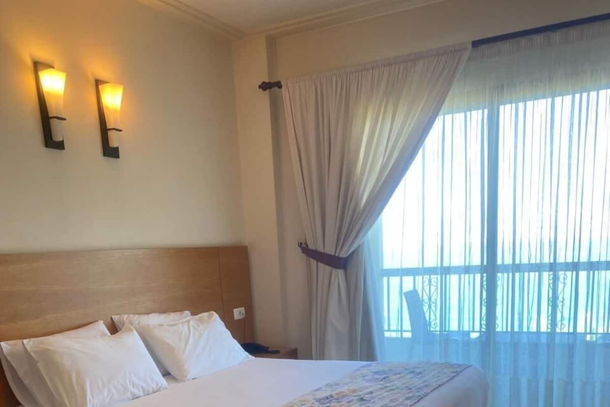 Une chambre d’hôtel avec un grand lit, une tête de lit en bois, deux lampes fixées au mur, une fenêtre avec des rideaux légers et une vue sur l’extérieur.