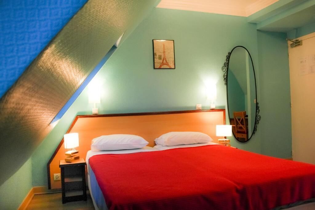A bedroom with a sloped ceiling, a bed with white pillows, a red blanket, a nightstand with a lamp, and a framed picture of the Eiffel Tower.