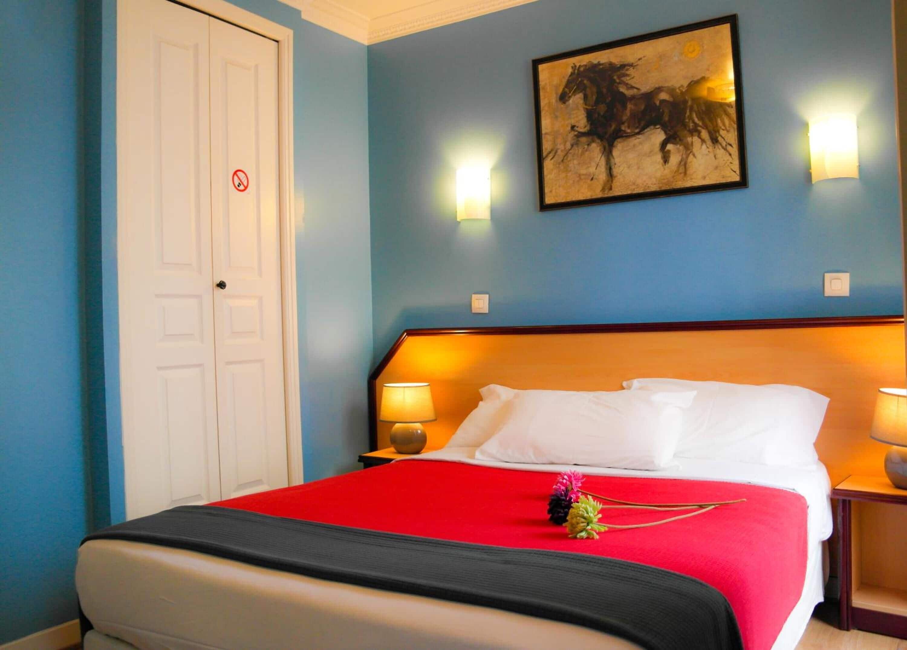 Habitación de hotel con una cama grande, una colcha roja, dos lámparas de noche, un cuadro de un caballo en la pared y una puerta blanca con un cartel de prohibido fumar.
