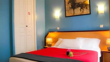 Habitación de hotel con una cama grande, una colcha roja, dos lámparas de noche, un cuadro de un caballo en la pared y una puerta blanca con un cartel de prohibido fumar.