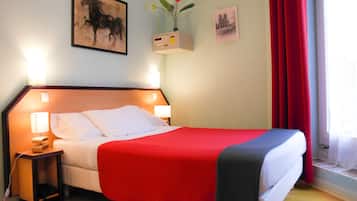 Una cama bien hecha con una colcha roja y gris, una mesita de noche con una lámpara, y una estantería con un jarrón de flores.