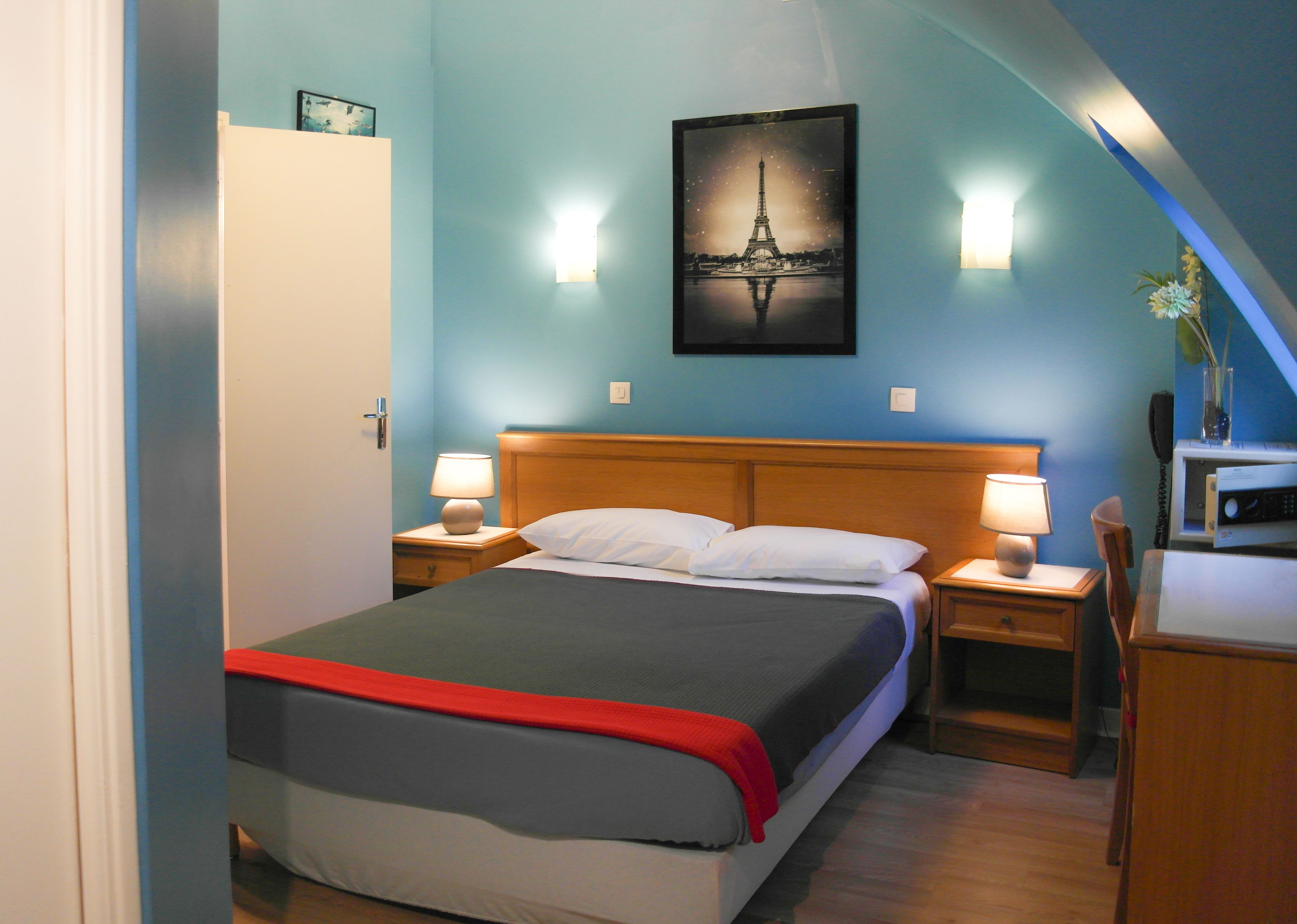 A hotel room with a bed, bedside lamps, a nightstand, a desk, and a picture of the Eiffel Tower on the wall.