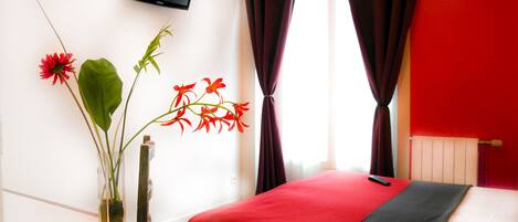 A hotel room with a bed, a small table with a vase of flowers, a television on the wall, and red curtains.