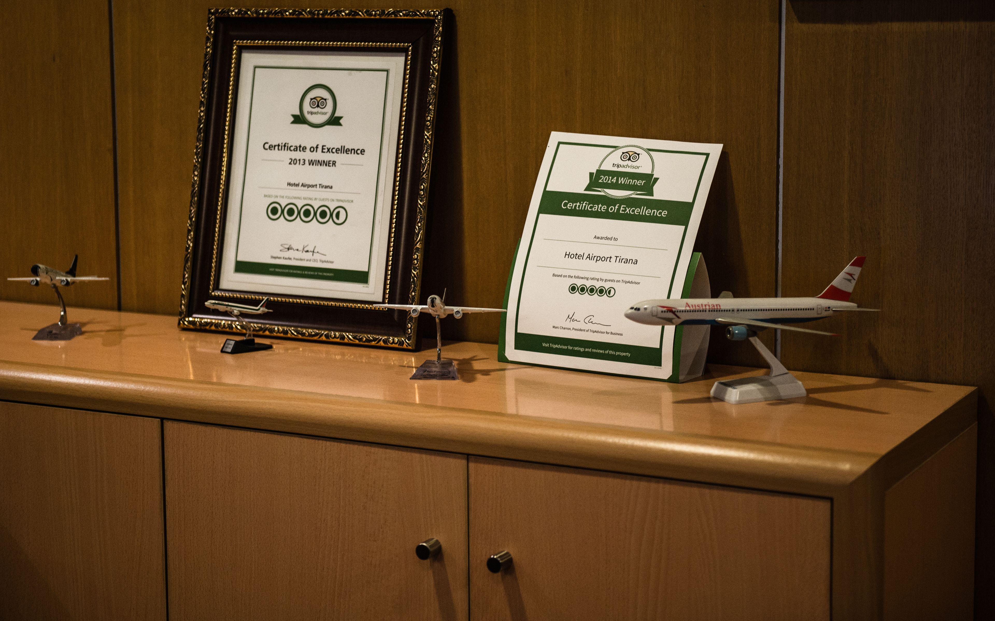Two framed certificates of excellence for Hotel Airport Tirana, one from 2013 and one from 2014, displayed on a wooden counter with a model airplane.
