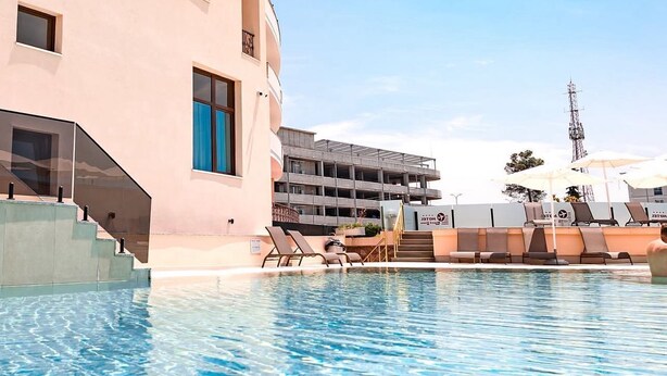 Une piscine d’hôtel avec une eau d’un bleu limpide, un escalier menant au bâtiment et une vue sur les environs, notamment un parking et une tour de communication.