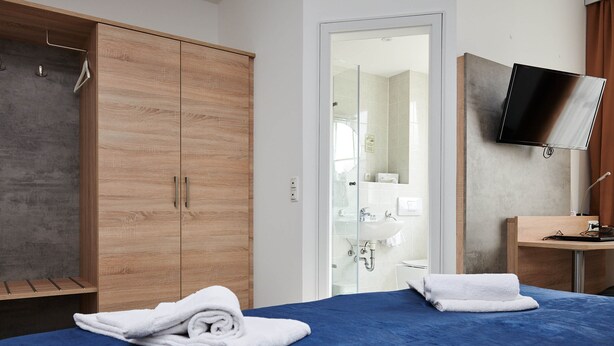 Habitación de hotel con cama, armario de madera, baño con ducha de vidrio y televisor montado.