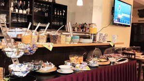 Montaje de bufé con diversas opciones de comida y bebidas, un televisor mostrando un noticiero y una zona de bar con botellas y vasos.