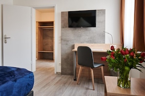 A hotel room with a bed, a desk with a chair, a TV mounted on the wall, and a vase of flowers on the desk. - Zur Allacher Muehle (Munich)
