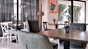 A modern dining area with a wooden table, white chairs, and a grey upholstered bench. A painting and a lamp are on the wall. - New Line Village Apartments (Sunny Beach)