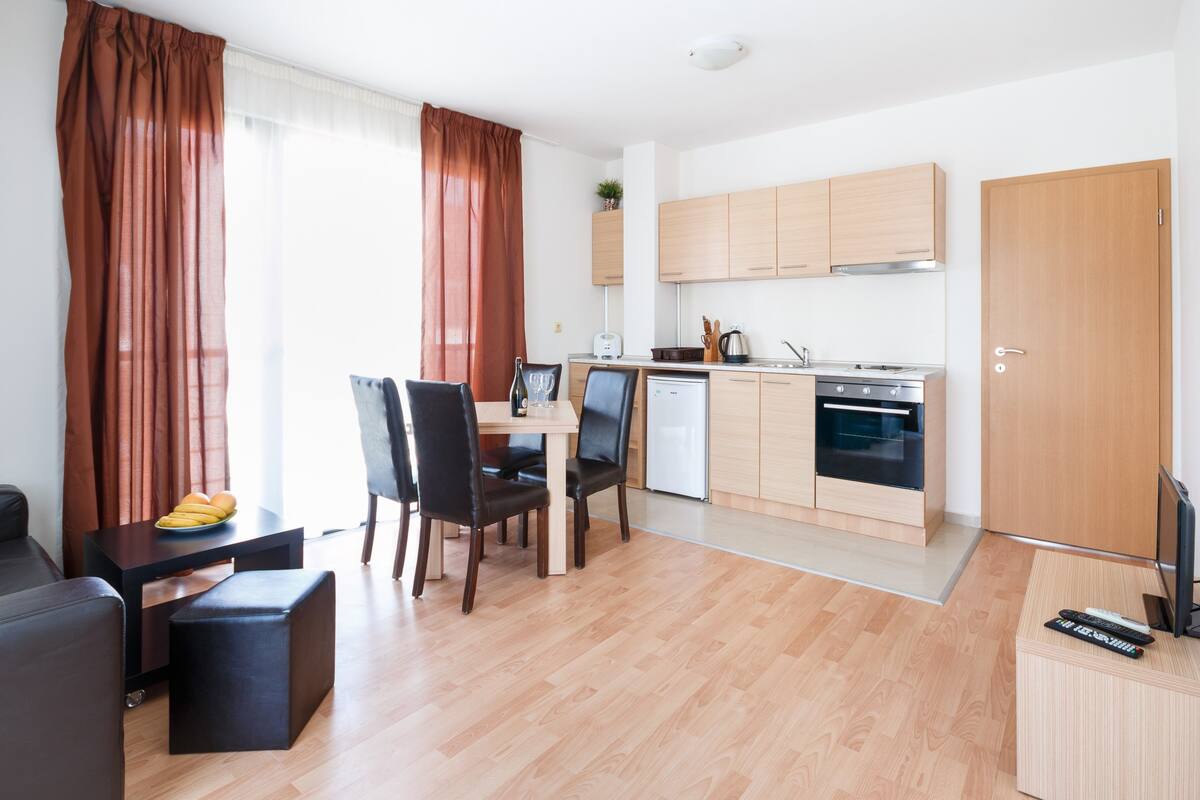 A modern kitchen with wooden cabinets, a dining area with a table and chairs, and a living room with a sofa and ottoman.