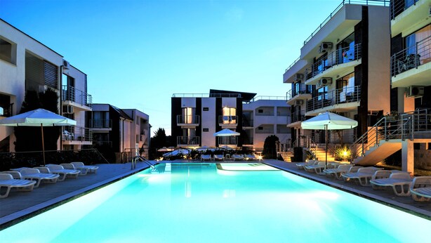 A swimming pool with lounge chairs and umbrellas, surrounded by modern buildings with balconies and outdoor seating. - New Line Village Apartments (Sunny Beach)
