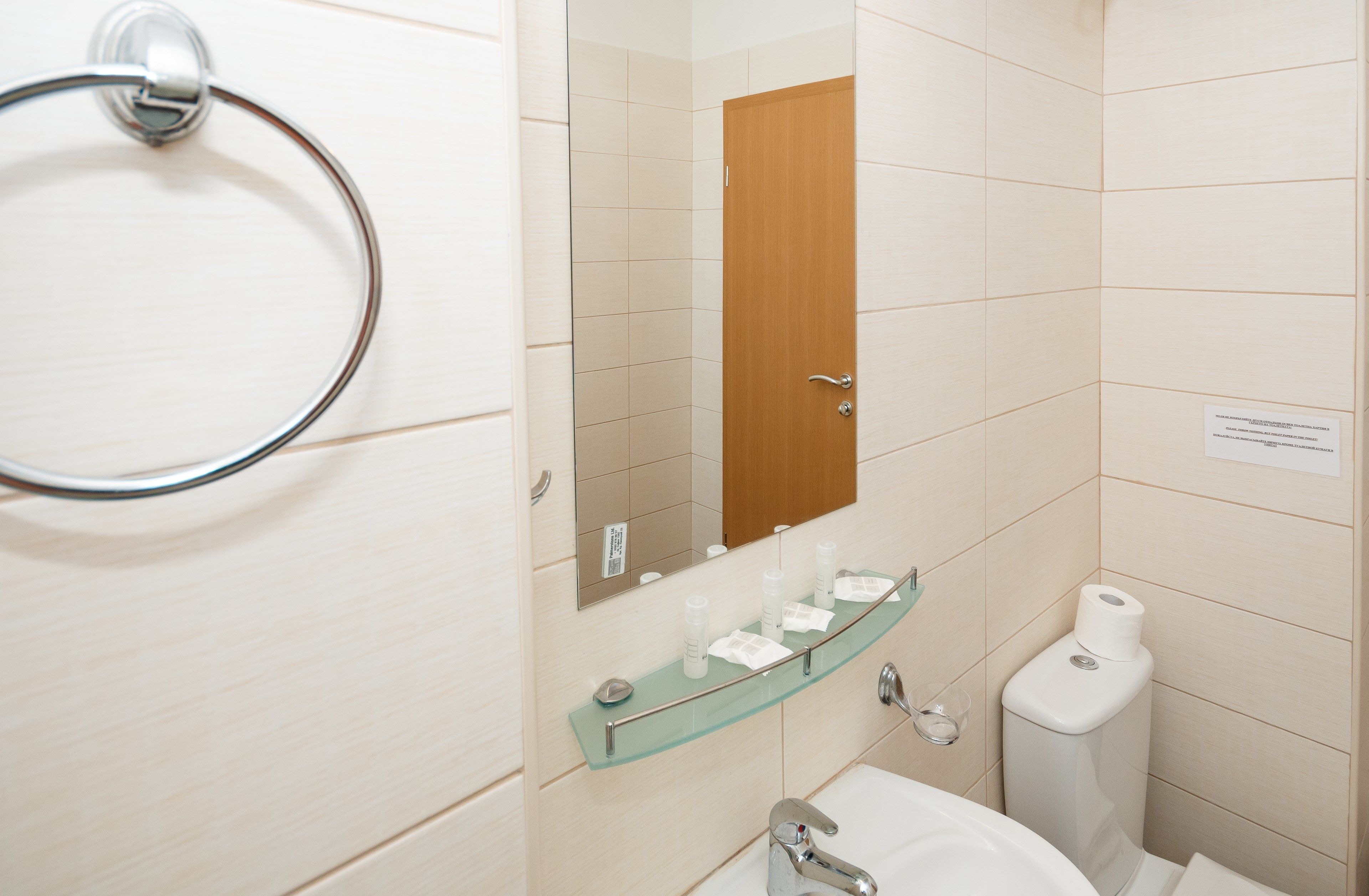 A bathroom with a glass shelf holding toiletries, a toilet, and a sink with a mirror above it.